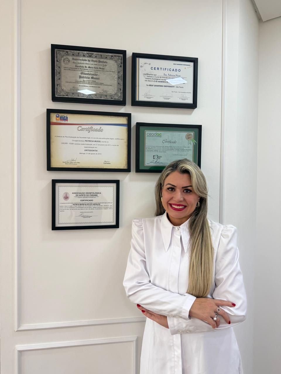 Dra. Patrícia Mussi vestida de branco, com os braços cruzados, diante de uma parede com certificados emoldurados.