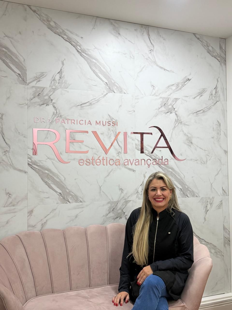 Dra. Patrícia Mussi sentada no sofá rosa da recepção, diante da parede de mármore com o nome Revita estética avançada.